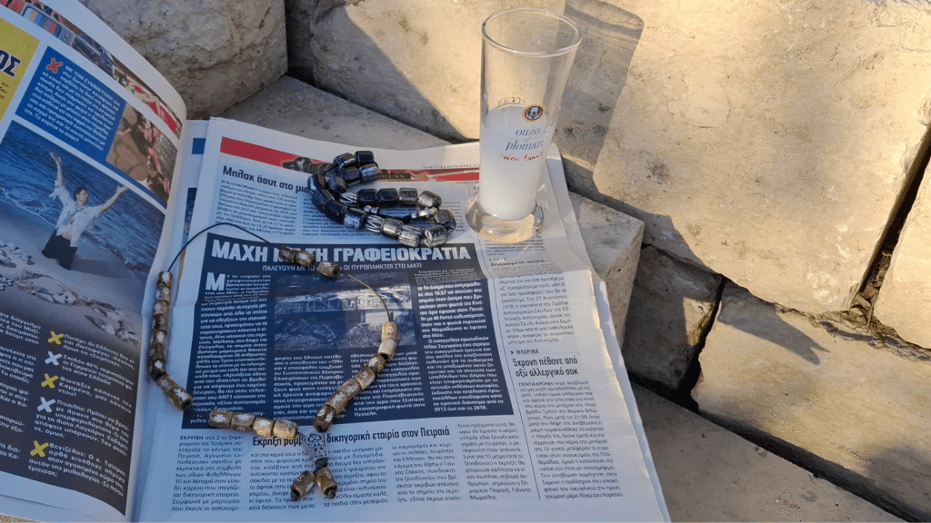 Zwei griechische Sorgenketten liegen neben einem Glas Ouzo auf einer Zeitung im Sonnenlicht. Das Komboloi ist ein Kulturgut.