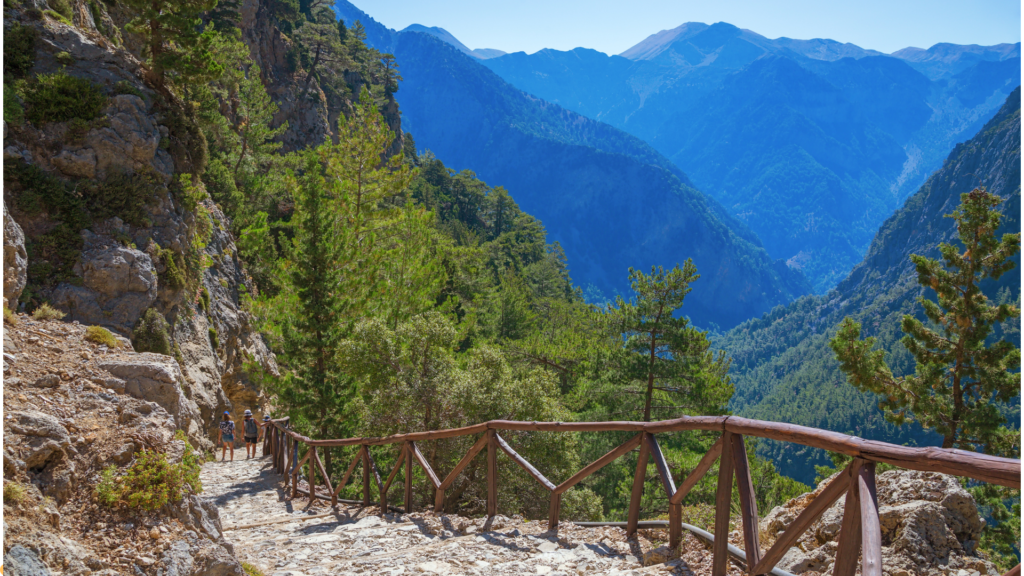 Kalderimia - Ein Kalderimi mit Stufen und Holzgeländer im Bergland von Kreta; sehr baumreich, im Hintergrund Berge, links vom Weg Felswände am sichtbaren Ende des Wegs vor einer Kurve zwei Wanderer.
